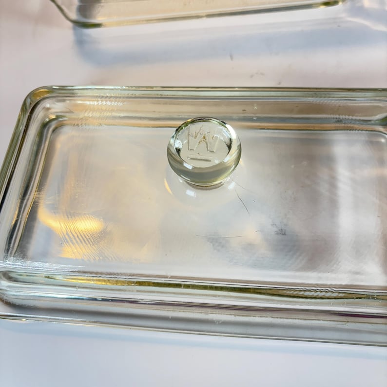May include: Clear glass rectangular dish with a rounded knob on the lid. The knob has an etched design. The dish is made of clear glass and has a simple, functional design. The dish is likely used for food storage or serving.