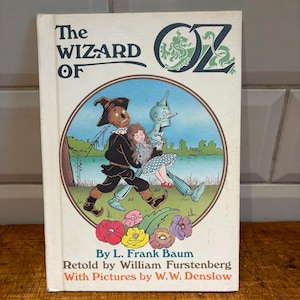 Puede incluir: Un libro antiguo de "El Mago de Oz". La portada presenta una ilustración colorida del Espantapájaros, Dorothy y el Hombre de Hojalata. El título está en letras grandes y en negrita, con el nombre del autor y otros detalles debajo.