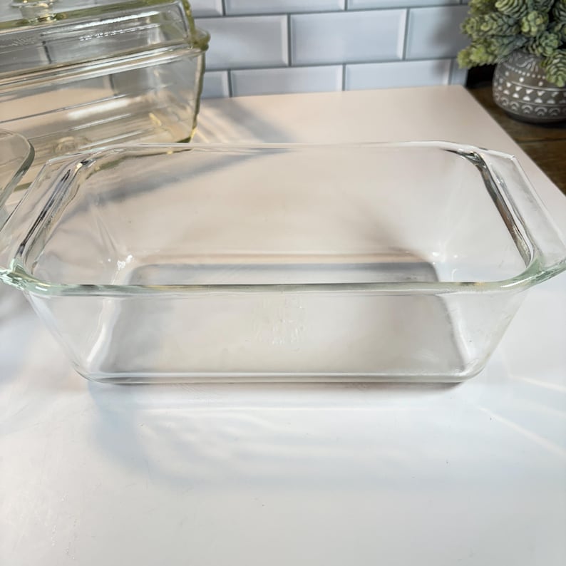 May include: A clear, rectangular glass baking dish with a slightly rounded bottom and handles on each side. Another glass dish with a lid is visible in the background. The dishes are on a white surface.