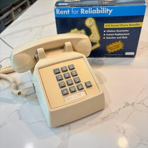 May include: A beige corded desk telephone with a push-button keypad and a coiled cord. The phone sits in front of its original blue and white GTE box, which features the text Rent for Reliability, GTE Rental Phone Benefits, and New includes a DATA JACK.