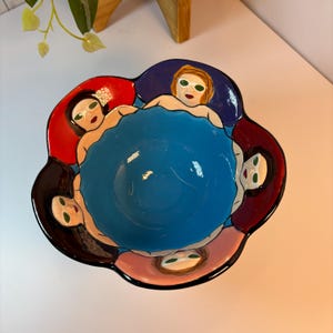 May include: A blue ceramic bowl with a flower-shaped rim. The bowl is decorated with five painted faces of women wearing sunglasses. The faces are surrounded by red, purple, pink, and black accents.