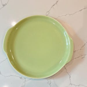 Op de afbeelding: Een ronde, lichtgroene keramische serveerschaal met twee gebogen handvatten. De schaal heeft een gladde, glanzende afwerking en is geschikt voor het serveren van hapjes of desserts. De kleur is een zacht, gedempt groen.