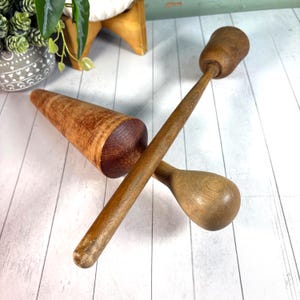 May include: Two wooden kitchen tools, a mortar and pestle, are displayed on a white surface. The mortar is a cone shape, and the pestle has a long handle with a rounded end. The wood has a warm, natural tone.