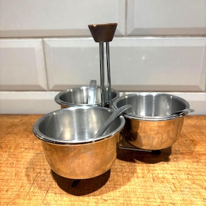 May include: A vintage condiment set with three stainless steel bowls, each with a small spoon. The set has a metal frame with a wooden handle. The bowls are arranged in a triangular shape.