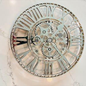 Può includere: Orologio da parete color argento a vista con numeri romani e ingranaggi visibili. Le lancette dell'orologio segnano circa l'1:10. L'orologio ha una finitura metallica ed è di forma circolare. È un oggetto decorativo.