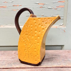 Puede incluir: Una jarra de cerámica amarilla con un asa marrón. La jarra tiene una superficie texturizada y una forma curva única.