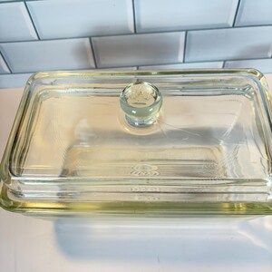 May include: A clear, rectangular glass butter dish with a matching lid. The lid features a round, clear knob with an embossed design. The dish is sitting on a white surface.