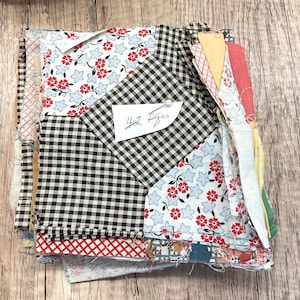 May include: A stack of fabric squares in various patterns and colors, including black and white gingham, floral prints with red flowers, and geometric designs. A small white tag with handwritten text is pinned to the fabric. The text on the tag reads "4x Pages".