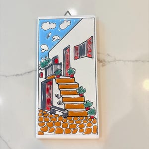 Puede incluir: Un azulejo rectangular de cerámica con una ilustración pintada a mano de un edificio blanco con escaleras naranjas, contraventanas rojas y plantas verdes en macetas. La escena incluye un cielo azul con nubes y un suelo de adoquines naranja.