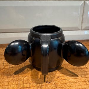 May include: Black novelty mug shaped like Mickey Mouse's head, featuring large, rounded ears on either side. The mug has a handle and a wide opening, perfect for beverages. The glossy finish gives it a classic, cartoonish look.