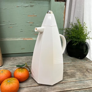 May include: White ceramic thermal carafe with a geometric design and a spout. The carafe has a silver top and a handle. The carafe is sitting on a wooden surface.