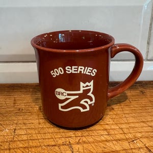 May include: A maroon ceramic mug with a curved handle. The mug features the text "500 SERIES" in white, along with a stylized logo and the letters "BRC". The mug is sitting on a wooden surface.