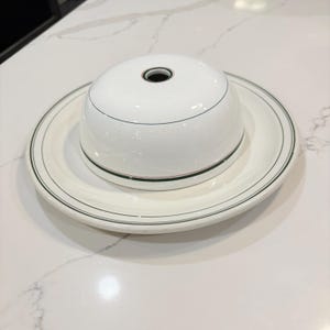 May include: A white ceramic plate and matching dome cover featuring a central hole. Both pieces are decorated with thin, concentric dark green and red circular lines. The dome sits centered on the plate, creating a cohesive serving set.
