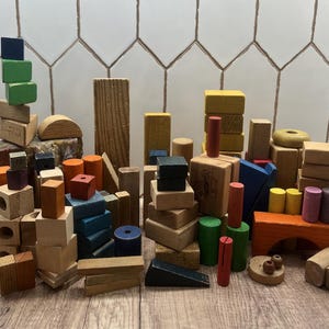 May include: A collection of wooden building blocks in various shapes and colors, including red, blue, green, and natural wood tones. The blocks are arranged on a wooden surface, showcasing a variety of geometric forms.