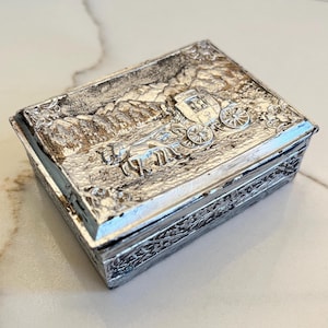 May include: A silver metal trinket box with an embossed design of a horse-drawn carriage. The box has a floral pattern around the edges.