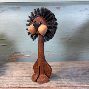 May include: A wooden lion figurine with a dark brown mane made of bristles. The lion has a dark brown body, a black nose, and two light brown cheek-like features. The figurine is standing on a wooden surface.
