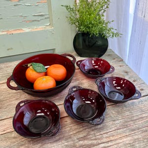 May include: Set of five ruby red glass bowls with handles. The bowls are arranged on a wooden surface with three oranges in the largest bowl.