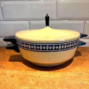 May include: A vintage yellow casserole dish with a black and white patterned band around the rim. The dish has a matching lid with a black finial and black handles on either side. The dish is sitting on a wooden surface.