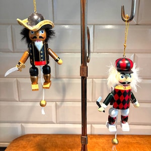 以下が含まれることがあります： 金色の紐で吊るされた、2つの木製の海賊をテーマにしたオーナメント。1つは黒い帽子とジャケットを着て、剣を持っています。もう1つは赤と黒の帽子とジャケットを着て、小さな武器を持っています。どちらも塗装されたディテールがあります。