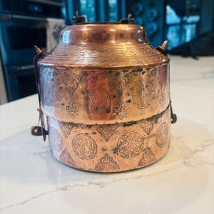 May include: A copper-colored, two-tiered container with intricate etched designs. The container has a cylindrical shape with a lid and a handle. The etched patterns include floral and geometric motifs, adding a decorative element to the piece.