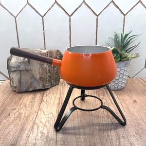 May include: A bright orange fondue pot with a smooth, rounded body and a long, dark brown wooden handle. The pot is supported by a black metal stand featuring three curved legs and a circular base ring for stability, designed for tabletop use.