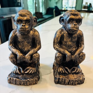 May include: Two bronze-colored monkey figurines in a seated position. The monkeys have a textured finish with gold highlights. They are sitting on a base that resembles a tree trunk. The figurines are displayed on a white countertop.
