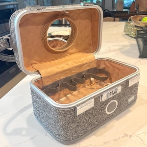 May include: A gray tweed hardshell vanity case with a silver metal frame and a combination lock. The interior is lined in tan fabric, featuring a circular mirror on the lid and a series of elastic loops for organizing items. The brand name Earhart is visible.