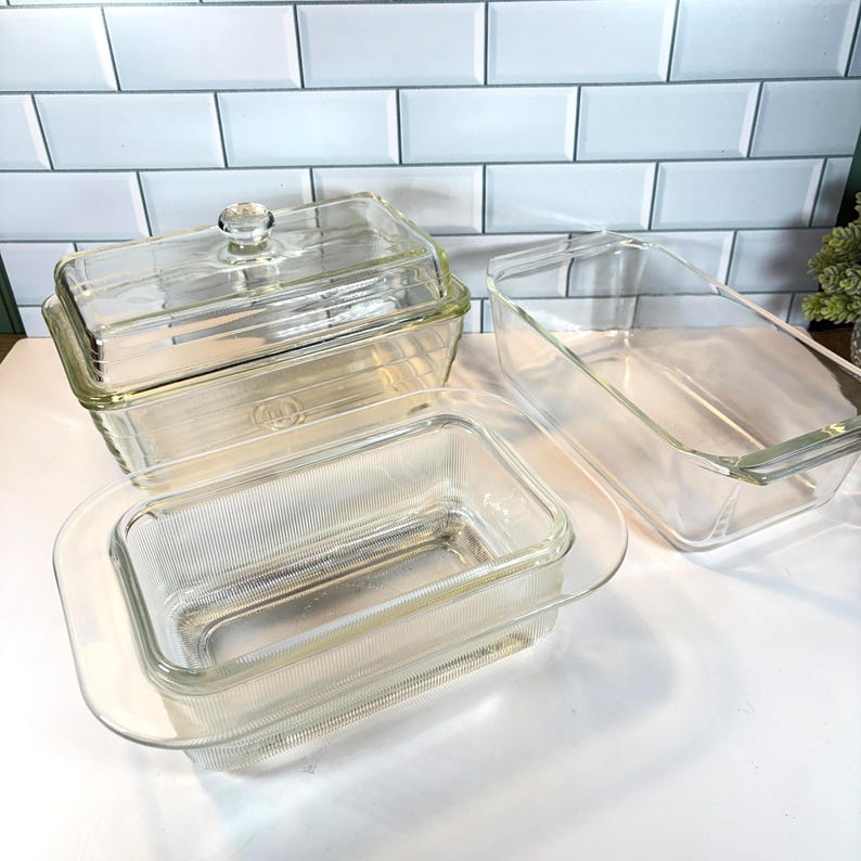 May include: A collection of clear glass baking dishes in various rectangular shapes. One dish has a lid with a round knob. The dishes are arranged on a white surface against a white tile background.