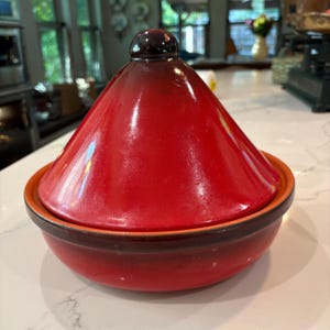 May include: A red and brown ceramic tagine pot with a conical lid and a round base. The lid has a dark brown knob. The pot is designed for slow cooking and is suitable for Moroccan cuisine.