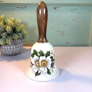 May include: A white ceramic bell with a brown wooden handle. The bell is decorated with white flowers, green leaves, and gold centers. The bell is sitting on a white surface.