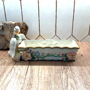 May include: A ceramic planter with a swan figurine on the left side. The rectangular planter is cream-colored with a gold trim and floral designs in pink, blue, and green. The swan is white with a yellow beak and a floral wreath.