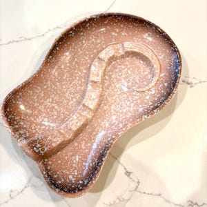 May include: A vintage ashtray with a unique shape and speckled design. The ashtray is primarily brown with white speckles and a dark brown rim. It features a curved design with a built-in rest for cigarettes.