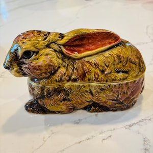 May include: A ceramic bunny figurine with a lid. The bunny is primarily brown with yellow and red accents. The ears are red inside. The figurine is likely a decorative item or a trinket box.