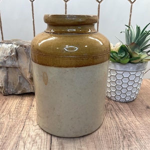 May include: A cylindrical stoneware crock featuring a two-tone design with a glossy brown glazed upper section and a matte cream-colored lower body. The vessel has a thick, rounded rim and a smooth, straight-sided profile with visible surface texture.