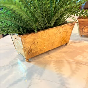 May include: A rectangular gold-toned metal planter with embossed swirling vine patterns on the sides. The planter features four small ball-shaped feet and is filled with a lush, vibrant green artificial fern plant with textured fronds.