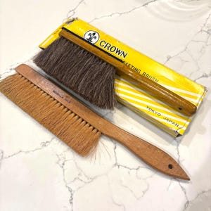 May include: Two wooden drafting brushes with natural bristles. One brush is labeled "MORILLA NO.3". A yellow box with the text "CROWN DRAFTING BRUSH TOKYO JAPAN" is also in the image. The brushes have wooden handles and are designed for art and drafting.