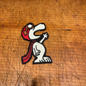 May include: Embroidered patch featuring Snoopy in a red aviator helmet and scarf. The patch is white, black, and red, with a detailed design of the iconic cartoon character. The patch is likely for clothing or accessories.