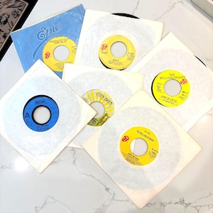 May include: A collection of six 7-inch vinyl records by The Rolling Stones, each housed in a white paper sleeve. The labels feature various colors including yellow, blue, and white, with printed text such as Start Me Up, Angie, and She's So Cold.