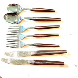 Puede incluir: Un juego de cubiertos de acero inoxidable con mangos marrones. El juego incluye dos cucharas, dos tenedores y dos cuchillos. Los mangos tienen un acento plateado. Los cubiertos están dispuestos sobre una superficie blanca.