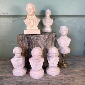 Può includere: Una collezione di piccoli busti bianco sporco di compositori famosi, tra cui Mozart, Schumann e Brahms. Ogni busto è dettagliato e montato su una base con il nome del compositore. I busti sono disposti su una superficie di pietra.
