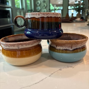 May include: Three ceramic bowls with handles, featuring a distinctive drip glaze pattern at the rim. The top bowl is dark blue, brown, and white. The bottom left bowl is tan and brown, while the bottom right bowl is light blue and brown.