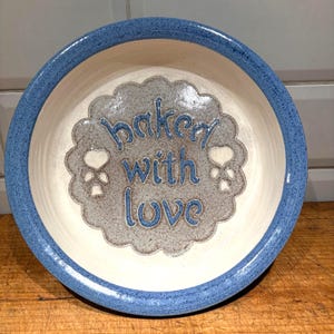 Puede incluir: Un cuenco de cerámica para mascotas con borde azul e interior crema. El centro presenta un diseño festoneado gris con las palabras "baked with love" en azul, junto con huellas de patas. El cuenco es para comida o agua.