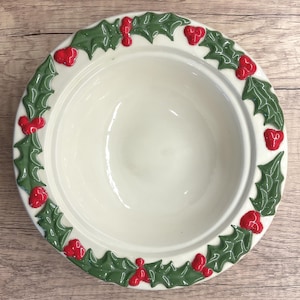 Könnte beinhalten: Eine weiße Keramikschale mit einem grünen Stechpalmen- und roten Beerenmuster am Rand. Die Schale hat eine glatte, glänzende Oberfläche und steht auf einer Holzoberfläche. Die Schale dient wahrscheinlich zum Servieren von Speisen.