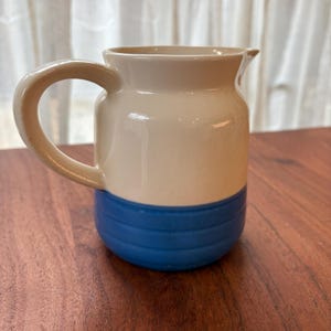 May include: A small cream-coloured ceramic creamer with a blue band around the bottom. The creamer has a handle and a rounded body.