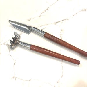 May include: Two small gardening tools with wooden handles and silver metal heads. One tool is a trowel, and the other is a small rake. The handles are a dark brown color, and the tools are set against a white marble background.