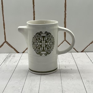 May include: White ceramic pitcher with a spout and handle. The pitcher features a green decorative design on the front. The base and rim have a green trim. The pitcher is on a white wooden surface.