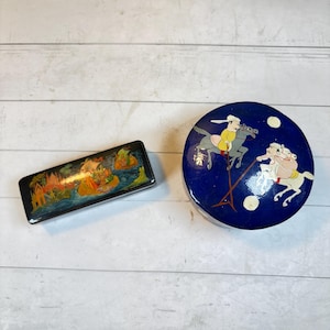 Puede incluir: Dos cajas decorativas. Una es rectangular y negra con una escena de paisaje colorida. La otra es redonda con un fondo azul y una escena pintada de dos figuras montando a caballo. Las cajas están sobre una superficie de madera blanca.