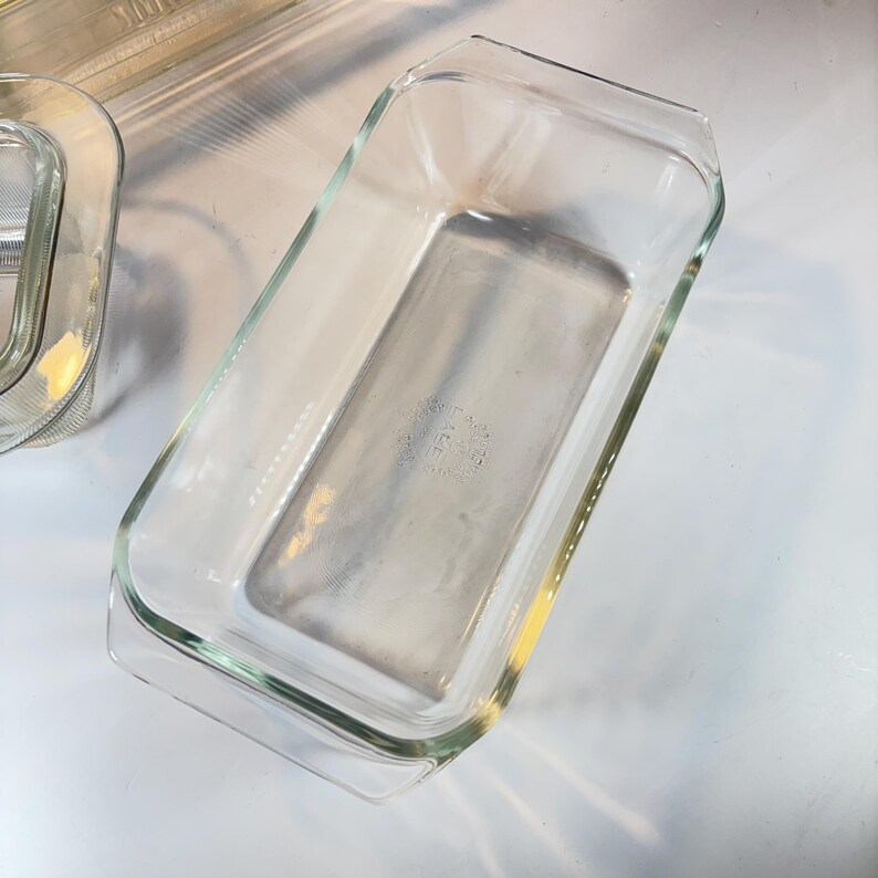 May include: Clear rectangular glass baking dish with slightly flared sides and a handle on each end. The dish is empty and appears to be vintage. Another similar dish is partially visible in the background.
