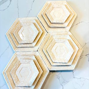 May include: A set of four off-white hexagonal plates with a textured, speckled surface. Each plate features a nested design with a central hexagon and layered edges. The plates are arranged on a white marble surface.