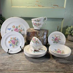 May include: A vintage white porcelain tea set with a floral pattern. The set includes plates, saucers, bowls, and teacups. The floral design features pink, yellow, and blue flowers. The set is arranged on a wooden surface.